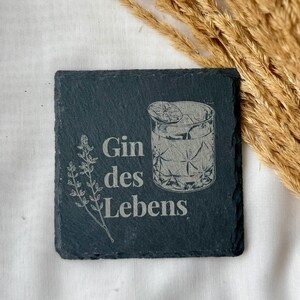 May include: Square black slate coaster with the words "Gin des Lebens" and a detailed illustration of a cocktail glass with ice and a lemon slice. A sprig of leaves is on the left.