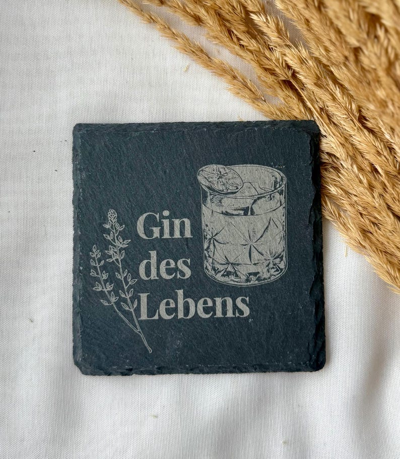 May include: Square black slate coaster with the words "Gin des Lebens" and a detailed illustration of a cocktail glass with ice and a lemon slice. A sprig of leaves is on the left.