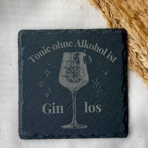 May include: A square, dark gray slate coaster with etched text and a cocktail glass design. The text reads "Tonic ohne Alkohol ist Gin los." The cocktail glass features a lemon slice. Decorative stars and hearts surround the glass.