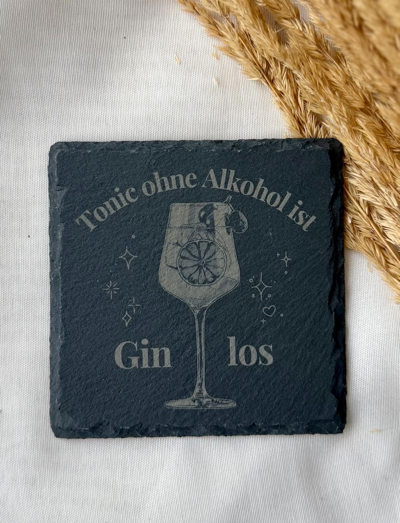 May include: A square, dark gray slate coaster with etched text and a cocktail glass design. The text reads "Tonic ohne Alkohol ist Gin los." The cocktail glass features a lemon slice. Decorative stars and hearts surround the glass.