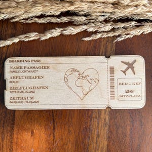May include: A wooden boarding pass with the text "BOARDING PASS", "NAME PASSAGIER", "FAMILIE LICHTWARDT", "ABFLUGHAFEN", "BERLIN", "ZIELFLUGHAFEN", "REYKJAV&Iacute;K, ISLAND", "ZEITRAUM", "04.09.2025-16.09.2025", "BER - KEF", "29F", "SITZPLATZ". The pass features a heart-shaped world map and a small airplane icon.