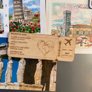 May include: A wooden boarding pass souvenir with a heart-shaped world map, airplane icon, and the text "BOARDING PASS", "NAME PASSAGIER", "FAMILIE LICHTWARDT", "ABFLUGHAFEN BERLIN", "ZIELFLUGHAFEN REYKJAVIK, ISLAND", "ZEITRAUM 04.09.2025-16.09.2025", "BER - KEF", "29F", and "SITZPLATZ".