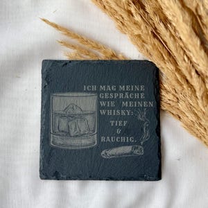 May include: A square, dark gray slate coaster with etched design. The design includes a whiskey glass with ice cubes, a cigar, and German text. The coaster is on a white surface with dried wheat stalks in the background.