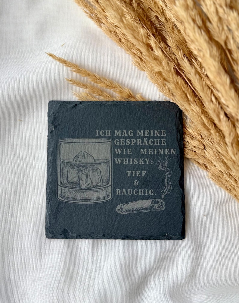 May include: A square, dark gray slate coaster with etched design. The design includes a whiskey glass with ice cubes, a cigar, and German text. The coaster is on a white surface with dried wheat stalks in the background.