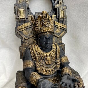 May include: A black and gold statue of a figure sitting on a throne. The figure is wearing a headdress and a necklace with a bird design. The throne is decorated with intricate carvings.