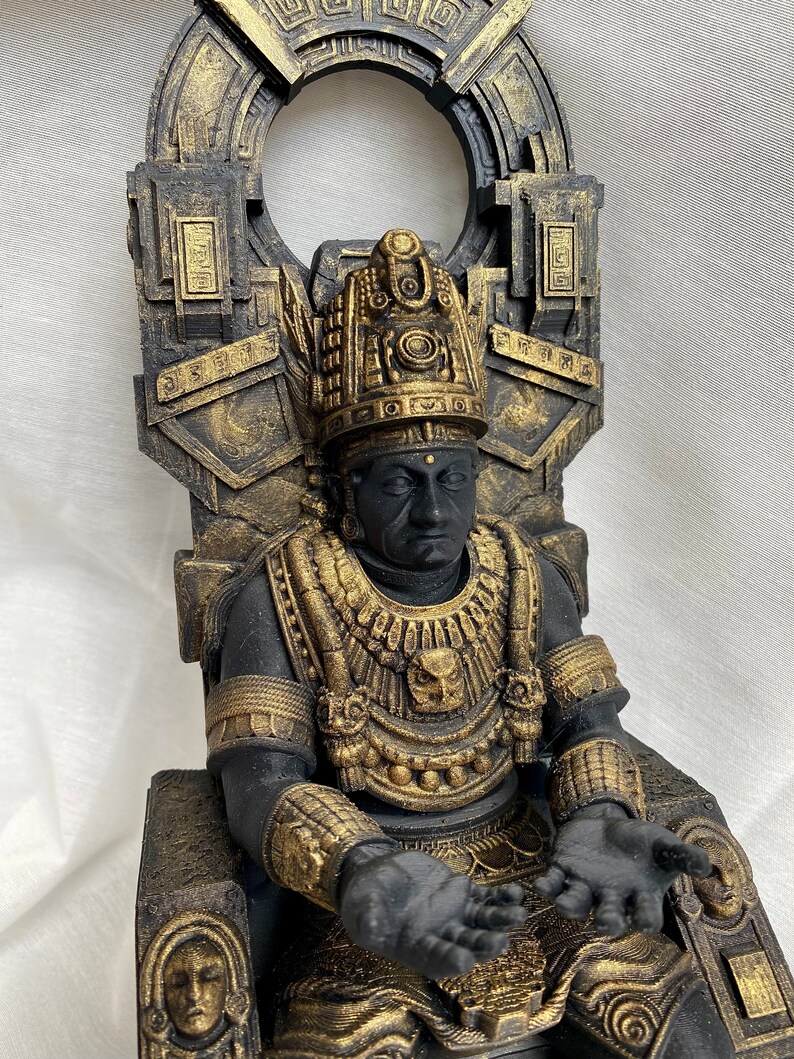 May include: A black and gold statue of a figure sitting on a throne. The figure is wearing a headdress and a necklace with a bird design. The throne is decorated with intricate carvings.