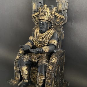 Könnte beinhalten: Eine schwarze und goldene Statue einer sitzenden Figur, die eine Krone und aufwendigen Schmuck trägt. Die Figur sitzt auf einem Thron mit kunstvollen Schnitzereien. Die Statue ist aus einem strapazierfähigen Material gefertigt und eine großartige Ergänzung für jede Inneneinrichtung.
