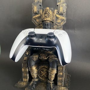 May include: A gold and black 3D printed stand shaped like a throne with a figure sitting on it. A white and black video game controller is resting on the throne.