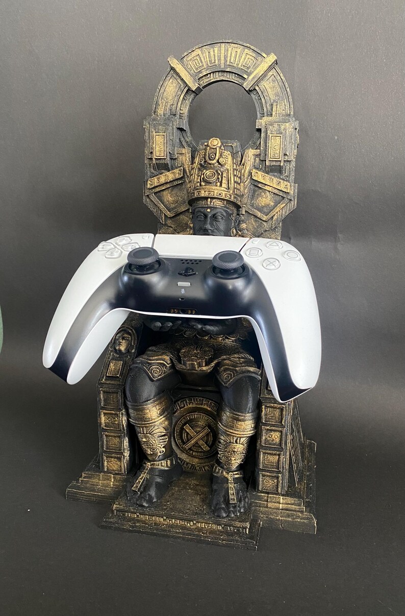 May include: A gold and black 3D printed stand shaped like a throne with a figure sitting on it. A white and black video game controller is resting on the throne.