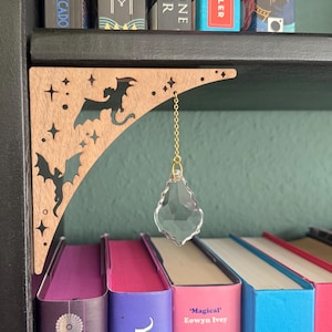 Può includere: Angolo di libreria in legno con intagli di pipistrelli e stelle, che tiene un ciondolo di cristallo trasparente su una catena d'oro. La mensola è sopra una fila di libri colorati, creando un arredamento stravagante di ispirazione gotica.