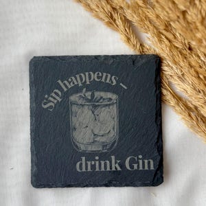 May include: Square black slate coaster with the phrase "Sip happens - drink Gin" etched on the surface. The coaster features an etched illustration of a cocktail glass filled with ice and a garnish. The background is a white surface with dried wheat.