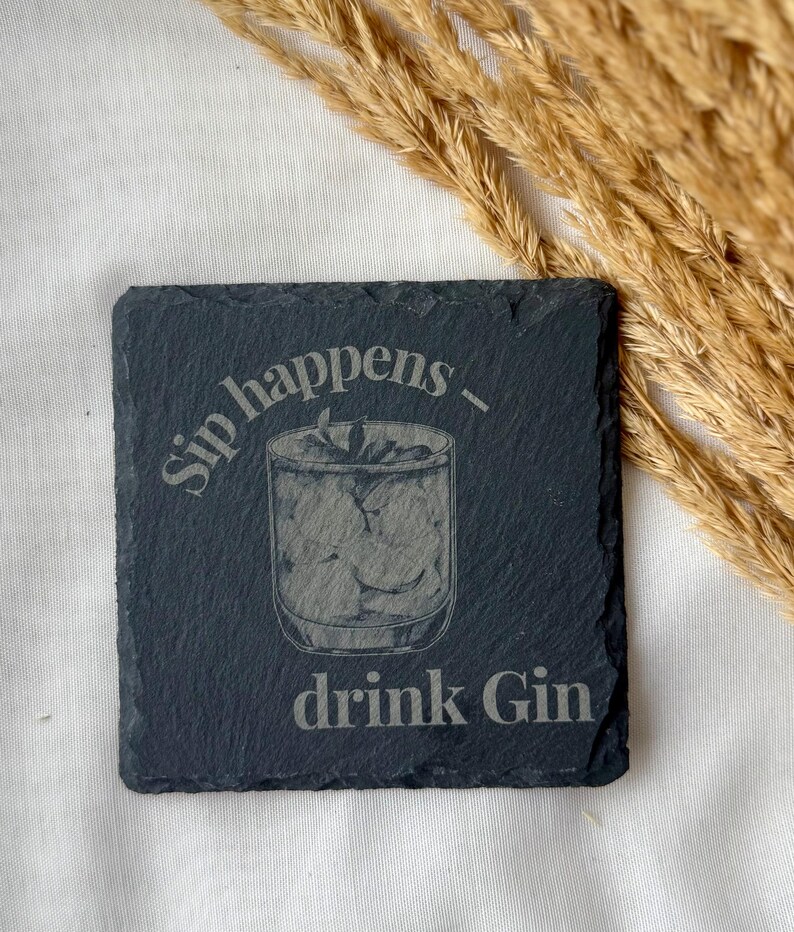 May include: Square black slate coaster with the phrase "Sip happens - drink Gin" etched on the surface. The coaster features an etched illustration of a cocktail glass filled with ice and a garnish. The background is a white surface with dried wheat.