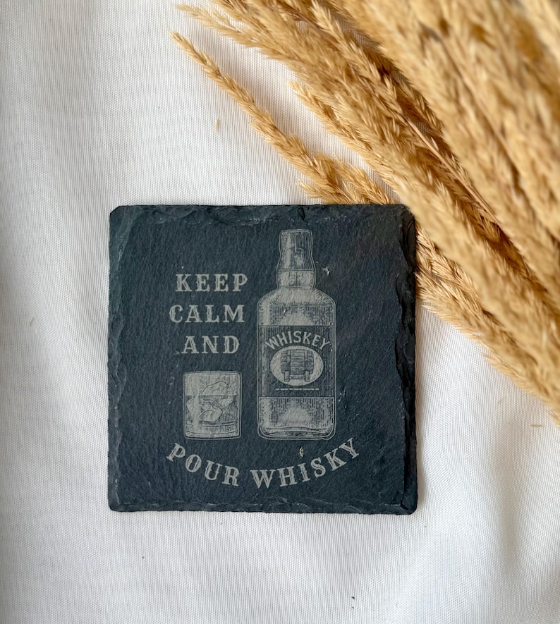 May include: A square, dark gray slate coaster with the words "KEEP CALM AND POUR WHISKEY" etched into the surface. The design includes an illustration of a whiskey bottle and a glass of whiskey.