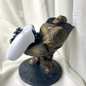 May include: A gold-painted figurine of a robot holding a white video game controller. The robot is standing on a black circular base with a decorative border.