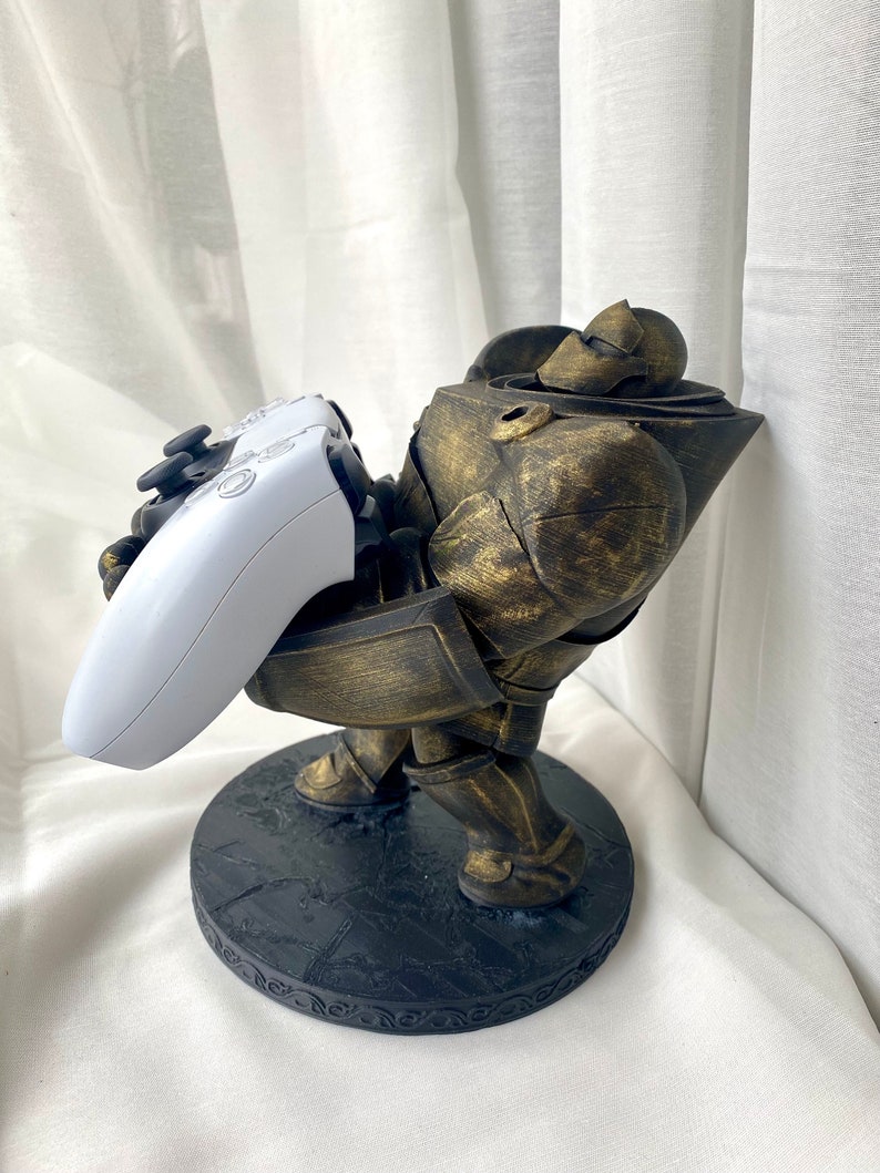May include: A gold-painted figurine of a robot holding a white video game controller. The robot is standing on a black circular base with a decorative border.