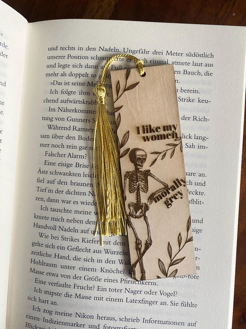 May include: A wooden bookmark with a skeleton illustration and the text "I like my women morally grey". The bookmark has a gold tassel and a floral design.