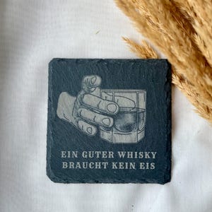 May include: Square, dark gray slate coaster with an etched design of a hand holding a whiskey glass. The text "EIN GUTER WHISKY BRAUCHT KEIN EIS" is etched below the image. The coaster is a bar accessory.