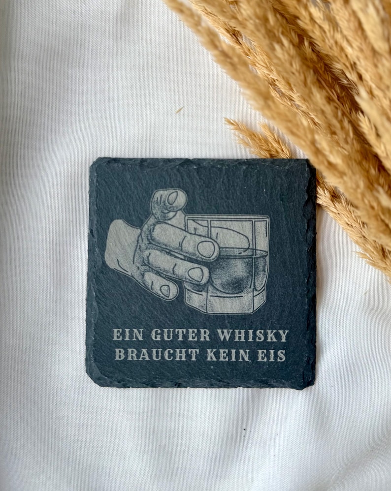 May include: Square, dark gray slate coaster with an etched design of a hand holding a whiskey glass. The text "EIN GUTER WHISKY BRAUCHT KEIN EIS" is etched below the image. The coaster is a bar accessory.