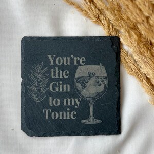 May include: A square, dark gray slate coaster with the text "You're the Gin to my Tonic" etched in a light gray color. The coaster features a drawing of a gin cocktail and a sprig of juniper.