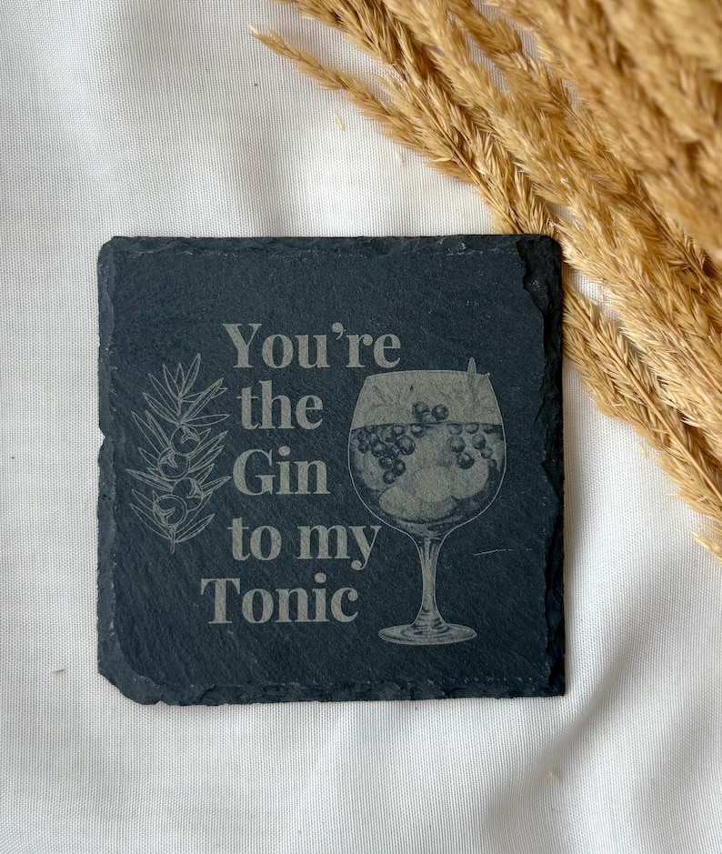 May include: A square, dark gray slate coaster with the text "You're the Gin to my Tonic" etched in a light gray color. The coaster features a drawing of a gin cocktail and a sprig of juniper.