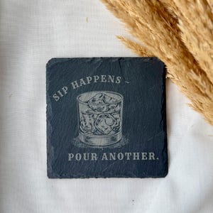 May include: Square, dark gray slate coaster with etched design. The design features a glass of ice cubes and the words "SIP HAPPENS - POUR ANOTHER." The coaster is a drink accessory.