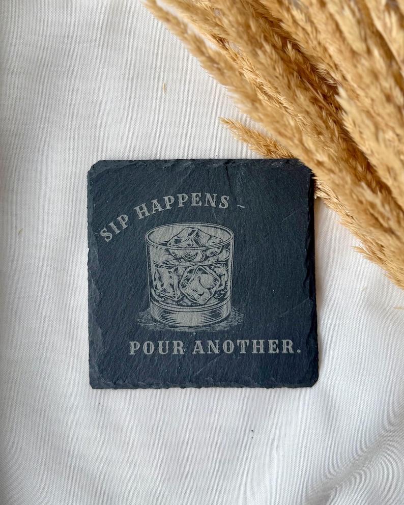 May include: Square, dark gray slate coaster with etched design. The design features a glass of ice cubes and the words "SIP HAPPENS - POUR ANOTHER." The coaster is a drink accessory.
