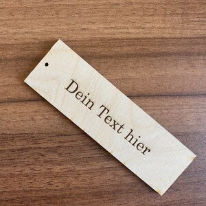 May include: A wooden tag with a hole in the top. The tag is light brown and has the text "Dein Text hier" engraved on it.