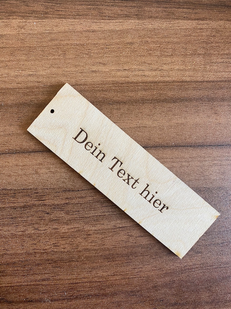 May include: A wooden tag with a hole in the top. The tag is light brown and has the text "Dein Text hier" engraved on it.