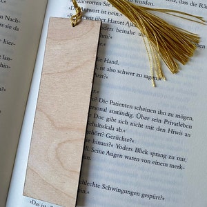 May include: A wooden bookmark with a gold tassel. The bookmark is blank and has a natural wood grain. The text on the page behind the bookmark reads "The patients seem to like him. He's personable.  He doesn't mess around with the interns on the pay scale."