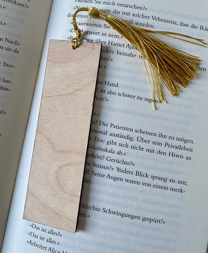 May include: A wooden bookmark with a gold tassel. The bookmark is blank and has a natural wood grain. The text on the page behind the bookmark reads "The patients seem to like him. He's personable.  He doesn't mess around with the interns on the pay scale."