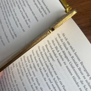 May include: A close-up of a gold-colored bookmark with a detailed design. The bookmark is resting on a page of a book with text in German. The text on the bookmark reads "Blutung stoppen!" which translates to "Stop the bleeding!" in English.