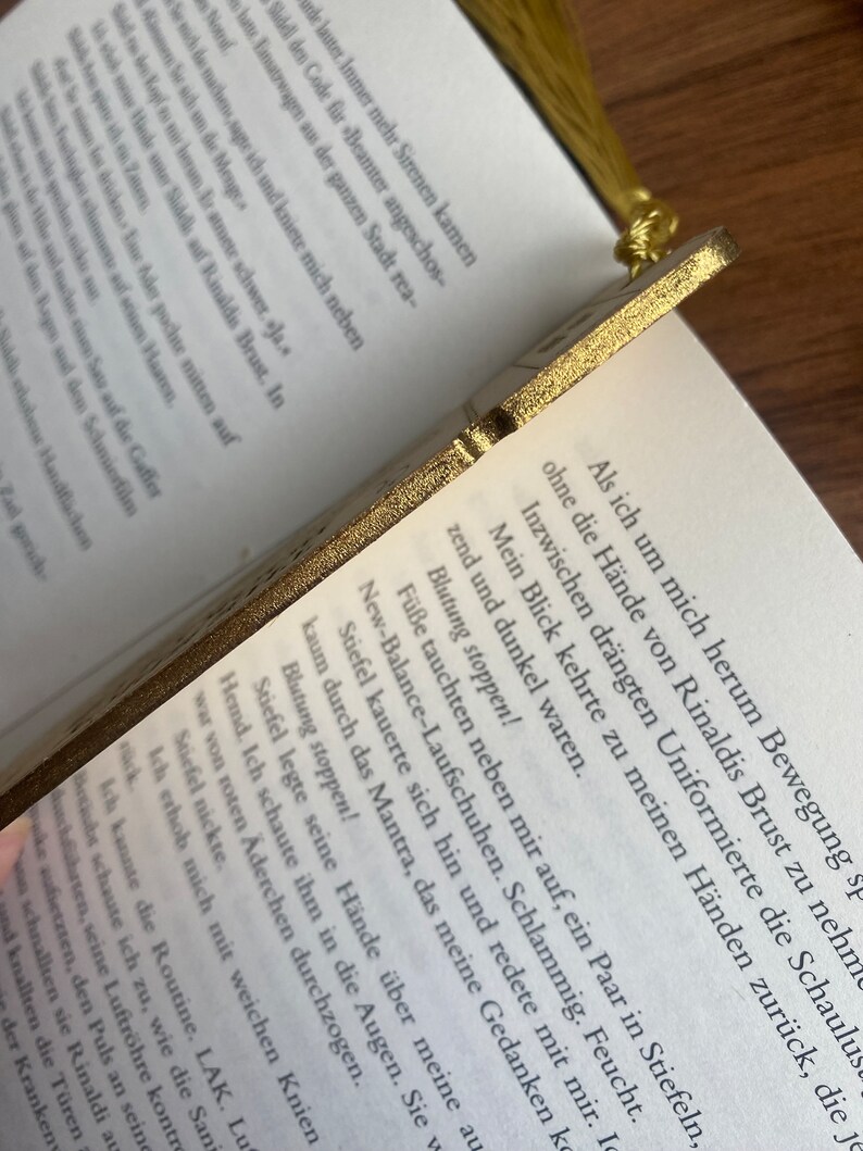 May include: A close-up of a gold-colored bookmark with a detailed design. The bookmark is resting on a page of a book with text in German. The text on the bookmark reads "Blutung stoppen!" which translates to "Stop the bleeding!" in English.