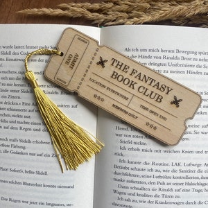 May include: A wooden bookmark shaped like a ticket with the text "The Fantasy Book Club" and stars. The bookmark has a yellow tassel attached to it. The bookmark is open to a page in a book with text about a character named Rinaldi.