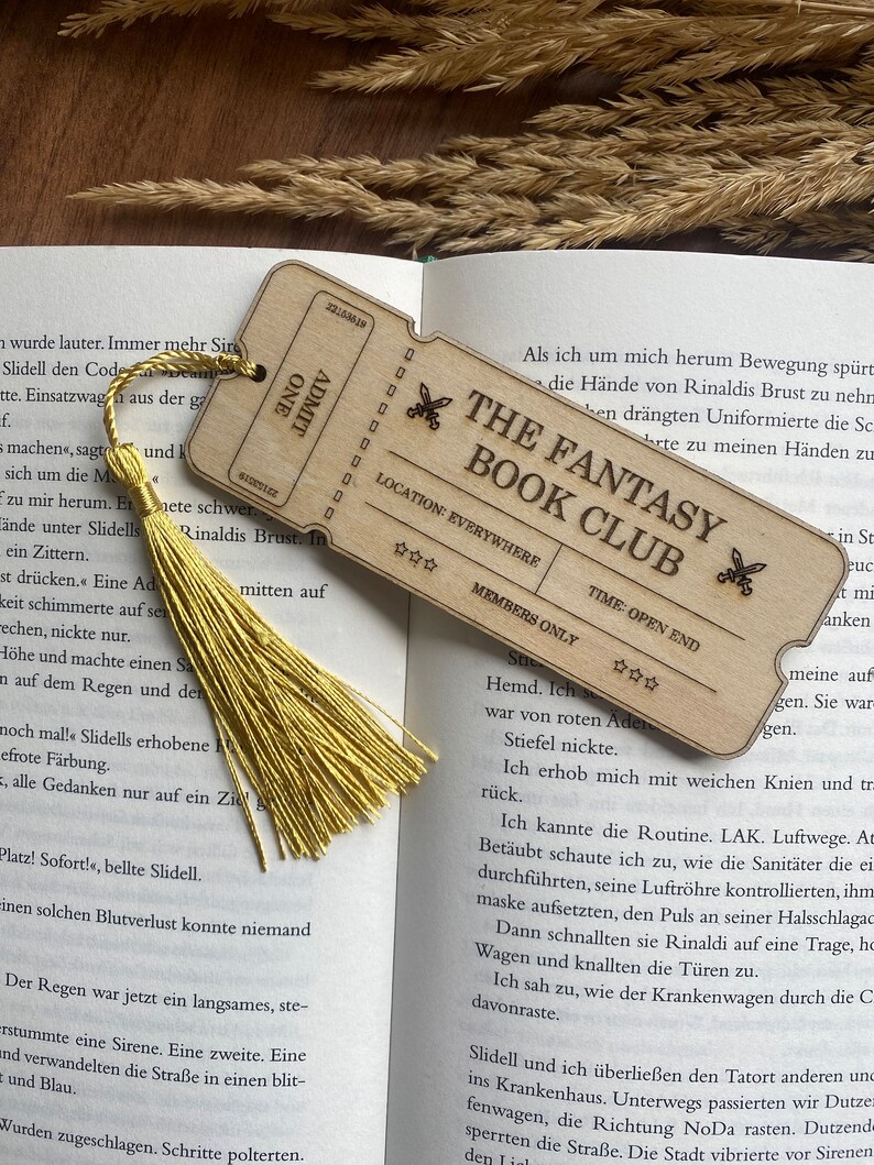 May include: A wooden bookmark shaped like a ticket with the text "The Fantasy Book Club" and stars. The bookmark has a yellow tassel attached to it. The bookmark is open to a page in a book with text about a character named Rinaldi.