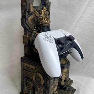 May include: A gold and black 3D printed statue of a figure holding a white PlayStation 5 controller. The statue is designed to hold a controller upright.