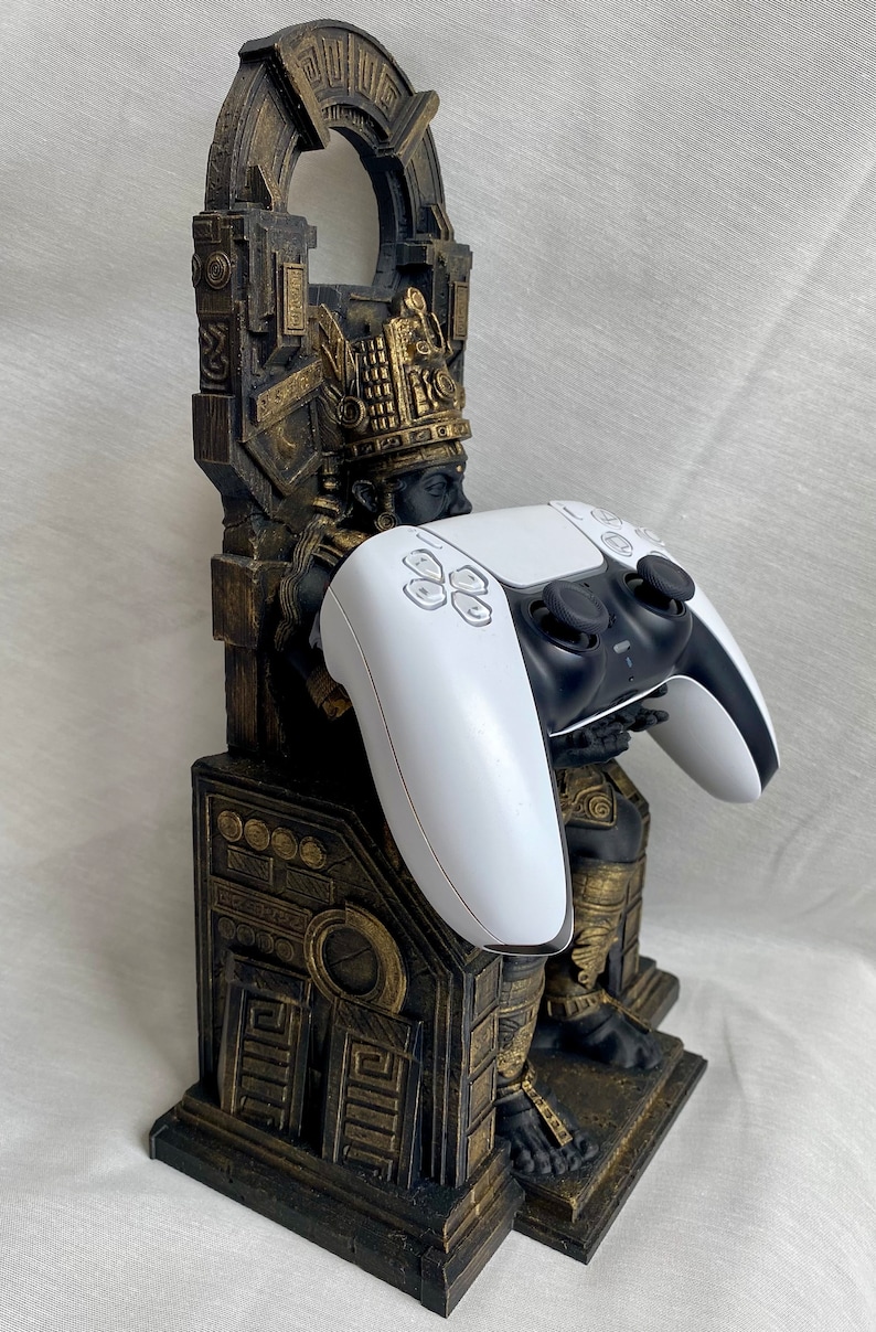 May include: A gold and black 3D printed statue of a figure holding a white PlayStation 5 controller. The statue is designed to hold a controller upright.