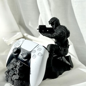 Könnte beinhalten: Ein schwarz-weißer PlayStation 5-Controller liegt auf einem schwarzen, strukturierten Sockel, mit einer schwarzen Figur eines Soldaten, der eine Waffe anvisiert. Der Controller ist weiß mit schwarzen Akzenten und Knöpfen. Die Figur ist detailliert und in Kampfpose.