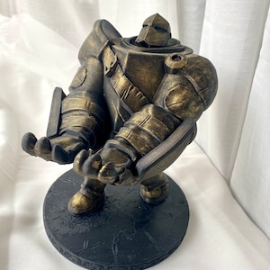May include: A gold-painted figurine of a robot with a round base. The robot is standing with its arms outstretched and has a textured surface.