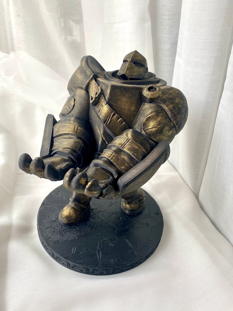 May include: A gold-painted figurine of a robot with a round base. The robot is standing with its arms outstretched and has a textured surface.