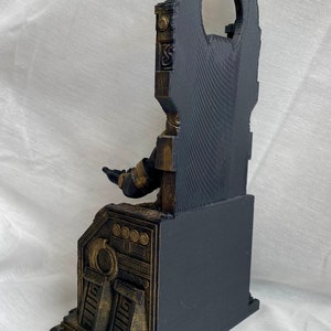 May include: A black and gold 3D printed statue of a figure sitting on a throne. The throne has intricate details and a large, circular opening in the back. The figure is wearing a black robe and has outstretched arms.