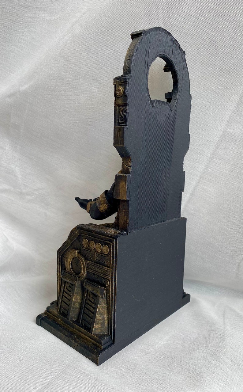 May include: A black and gold 3D printed statue of a figure sitting on a throne. The throne has intricate details and a large, circular opening in the back. The figure is wearing a black robe and has outstretched arms.