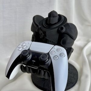 May include: A black and white video game controller held by a black figurine. The controller has a white face and black buttons. The figurine is a stylized, armored figure with a round base.
