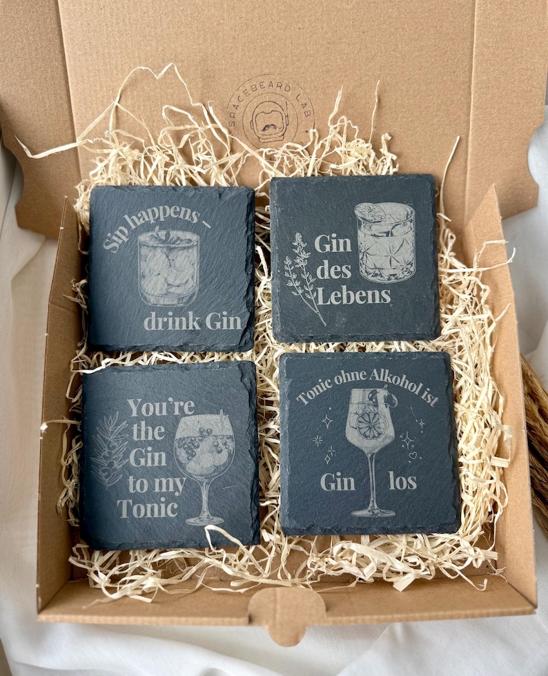 May include: A set of four square slate coasters with engraved gin-themed designs and phrases, including "Sip happens - drink Gin" and "You're the Gin to my Tonic." The coasters are presented in a cardboard box with straw.