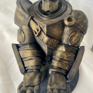 May include: A gold-painted 3D printed figurine of a robot wearing armor. The robot is sitting with its legs crossed and its arms folded. The figurine is detailed with textured surfaces and a weathered look.