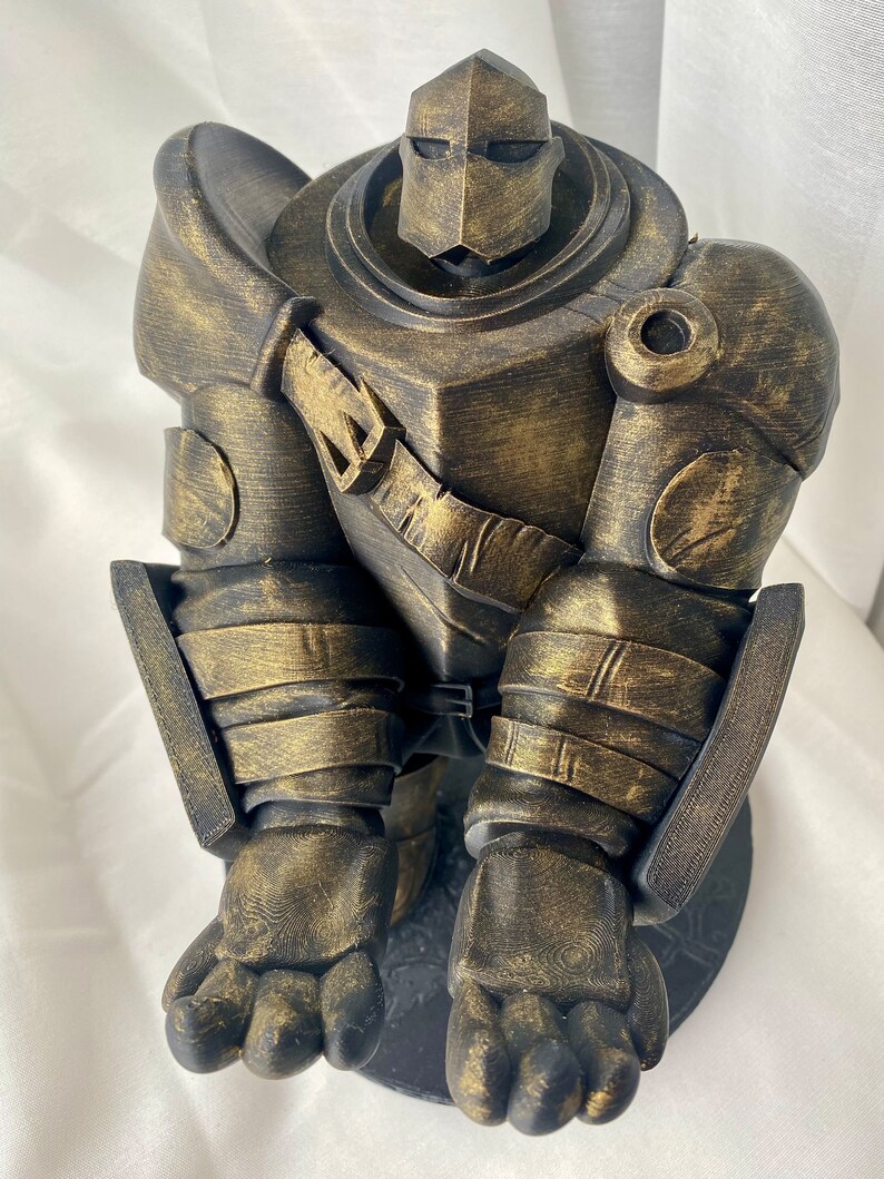 May include: A gold-painted 3D printed figurine of a robot wearing armor. The robot is sitting with its legs crossed and its arms folded. The figurine is detailed with textured surfaces and a weathered look.