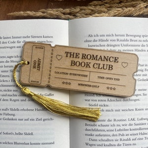 May include: A wooden bookmark with a gold tassel. The bookmark is shaped like a ticket and has the text "The Romance Book Club" printed on it. The bookmark also has a heart symbol and stars.