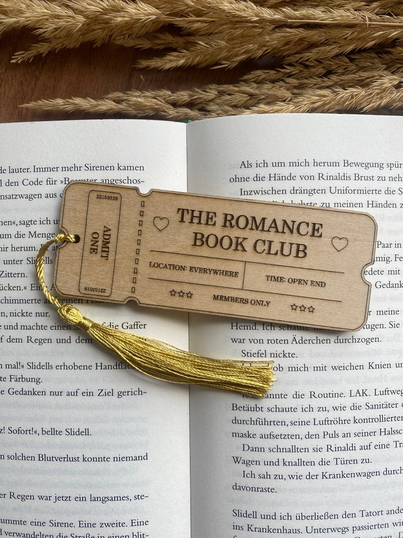 May include: A wooden bookmark with a gold tassel. The bookmark is shaped like a ticket and has the text "The Romance Book Club" printed on it. The bookmark also has a heart symbol and stars.
