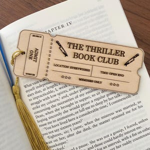 May include: A wooden bookmark shaped like a ticket for "The Thriller Book Club". The ticket has a red and white striped design with the words "The Thriller Book Club" in black text. The ticket also has the words "Location: Everywhere" and "Time: Open End" in black text. The bookmark has a gold tassel.