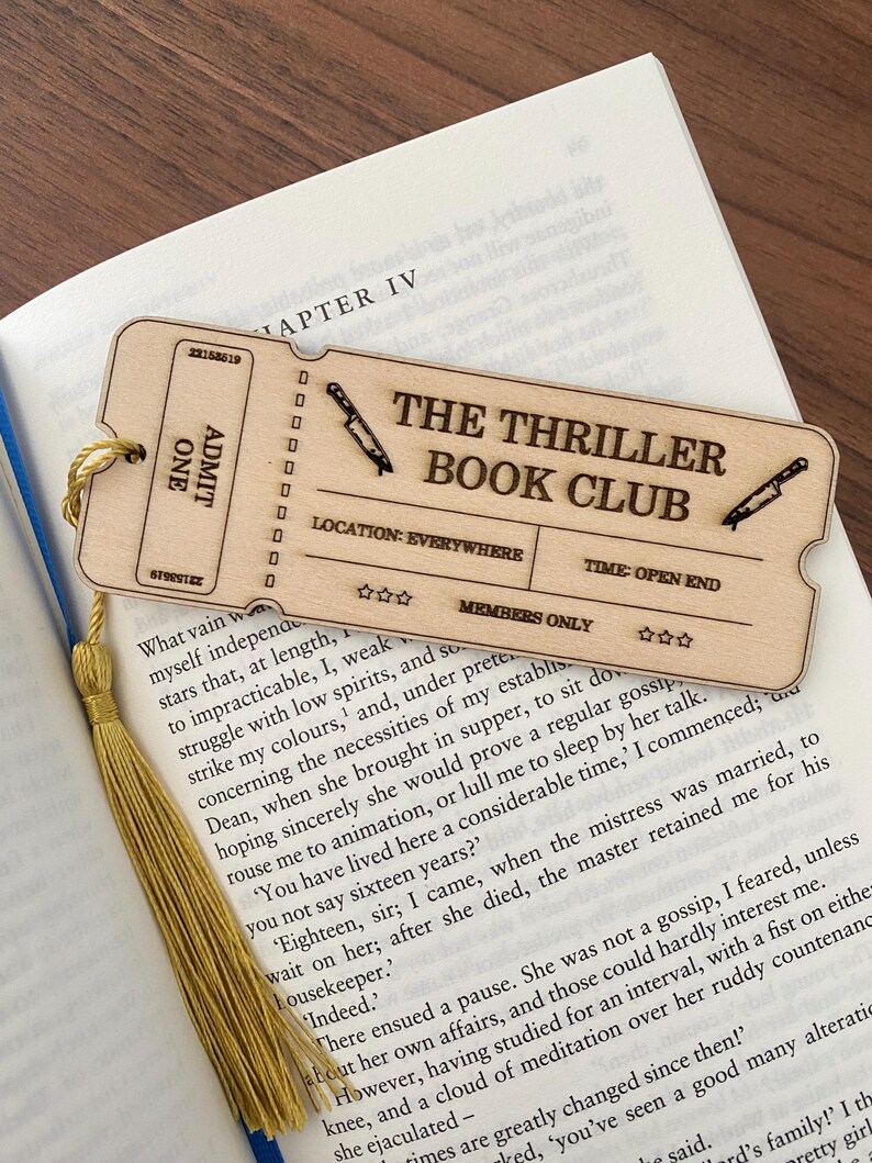 May include: A wooden bookmark shaped like a ticket for "The Thriller Book Club". The ticket has a red and white striped design with the words "The Thriller Book Club" in black text. The ticket also has the words "Location: Everywhere" and "Time: Open End" in black text. The bookmark has a gold tassel.