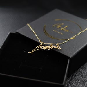 May include: A gold necklace with a dolphin pendant featuring the name "Angelina" in cursive script. The necklace is displayed in a black box with a white logo.